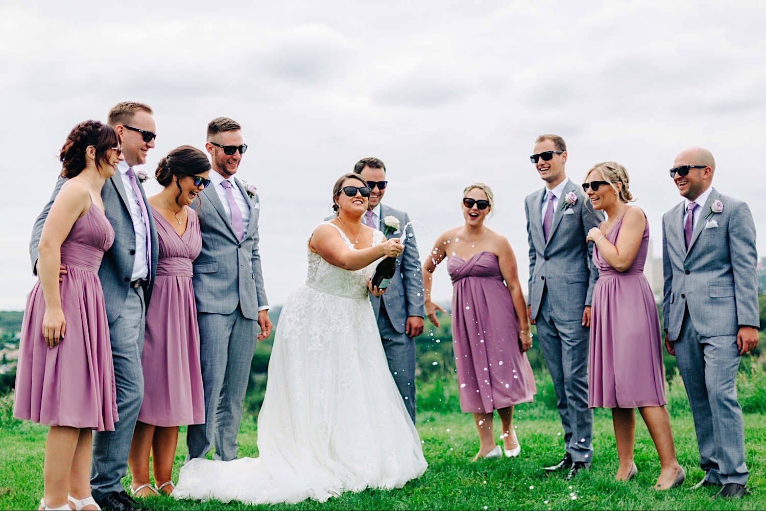 Bride popping a champagne bottle with bridesmaids in purple dresses and groomsmen in grey suits. Everyone is wearing sunglasses. Downtown skyline and overcast day.